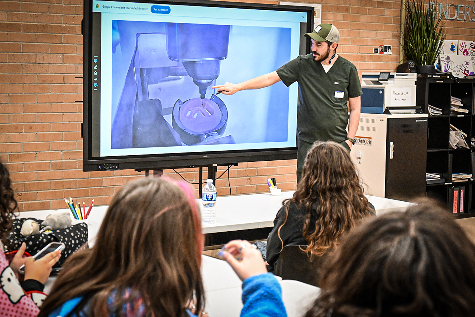 A man in scrubs and a baseball hat points to a screen with a dental drill