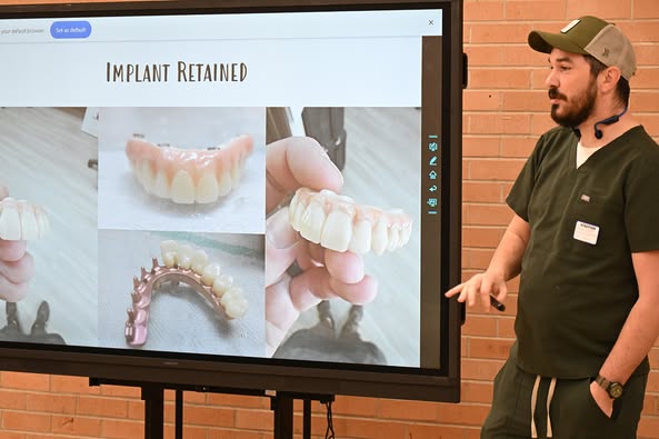 A man in scrubs and a baseball hat stands in front of a screen with dental implant examples