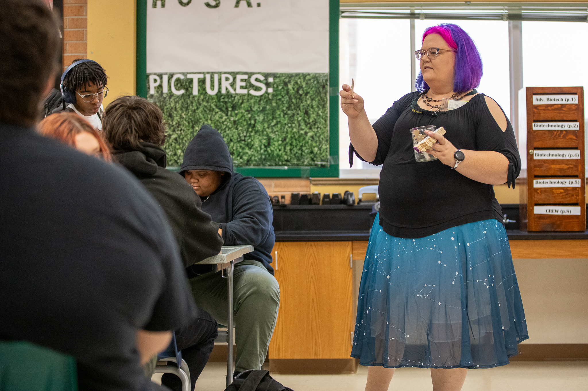 A teacher with bright purple hair holds up popsicle sticks in her classroom