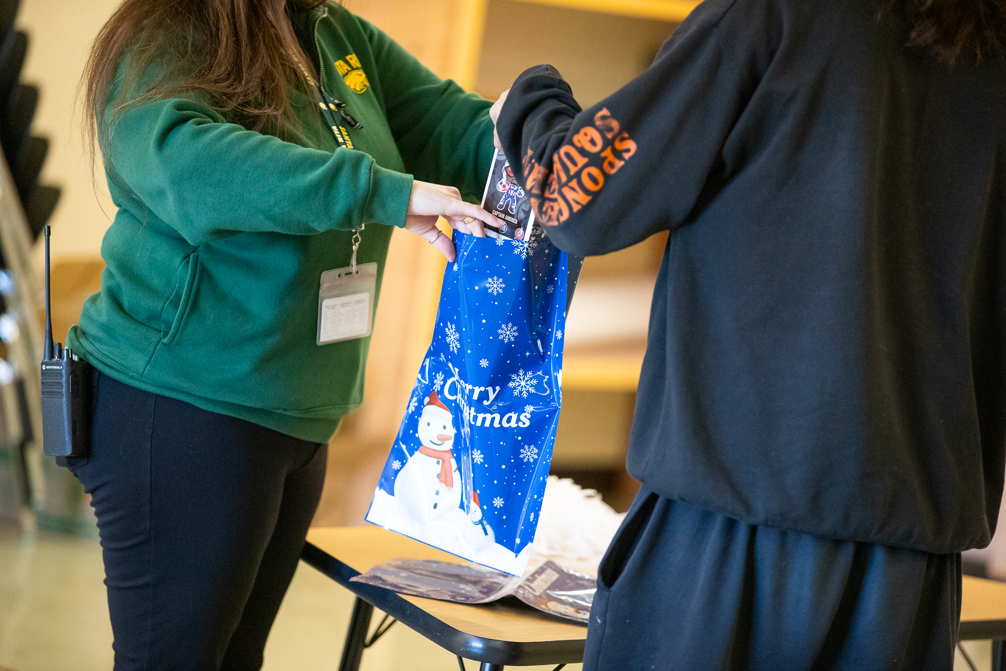 A woman in a green sweatshirt hands a student in a black hoodie a blue Merry Christmas gift bag with a snowman on it