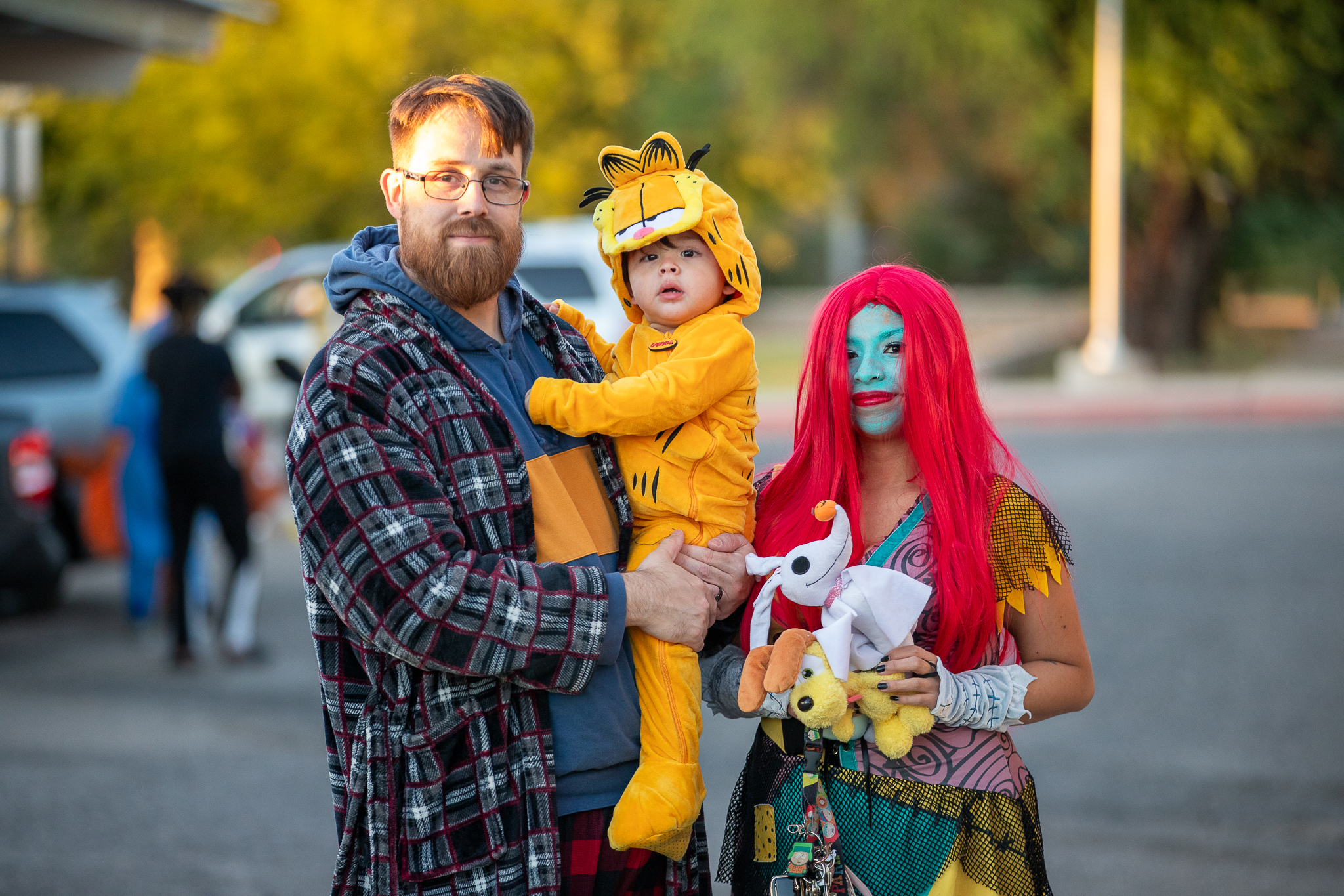 A dad holds his son in a Garfield costume, while the boy's son stands next to them in a Sally from Nightmare Before Christmas costume