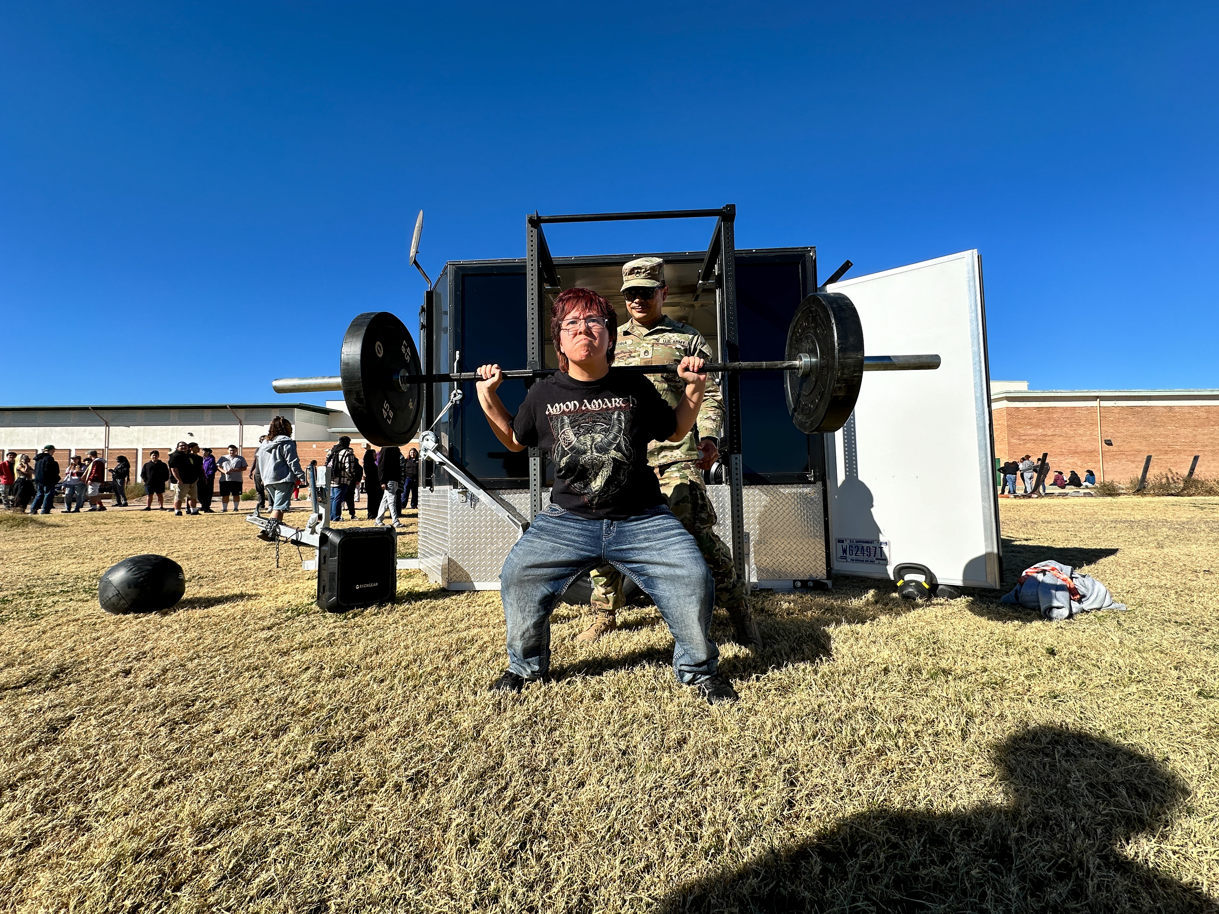 A student attempts a squat with a barbell while a man in an Army uniform looks on