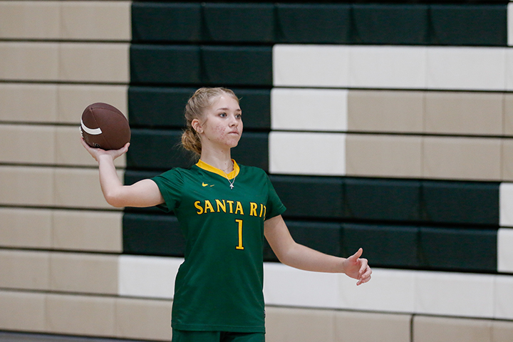 A teen girl with blonde hair wearing a green Santa Rita jersey throws a football inside the gym