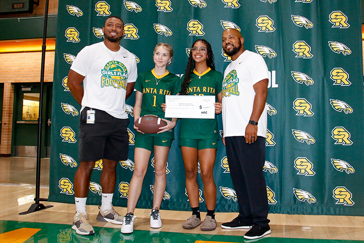 Two teen girls, center, hold a football and a check, with their coaches on either side of them