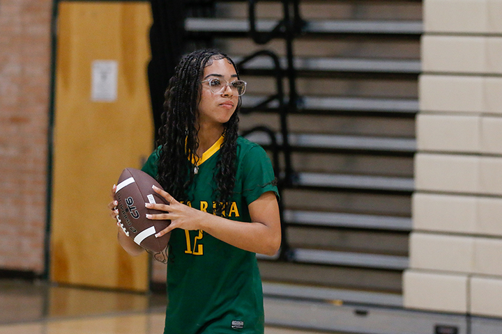 A teen girl with glasses and long dark curly hair wearing a green jersey gets ready to throw a football in the gym