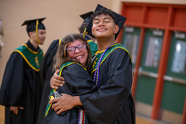 A teen girl and teen boy smile and hug each other in their graduation regalia